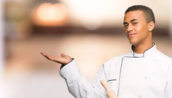 Young afro american chef man holding copyspace imaginary on the palm to insert an ad on unfocused background
