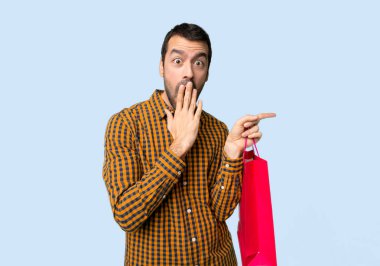 İzole mavi arka planda sürpriz bir yüz ile yan parmak işaret alışveriş çantaları ile adam