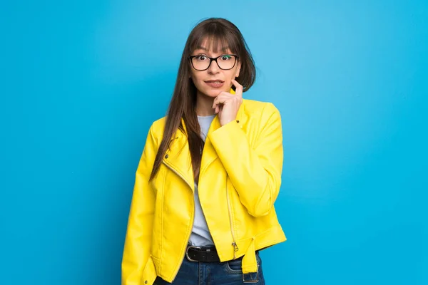 Young woman with yellow jacket on blue background having doubts while looking up