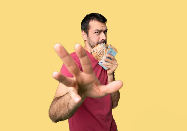 Man taking a lot of money is a little bit nervous and scared stretching hands to the front on isolated yellow background