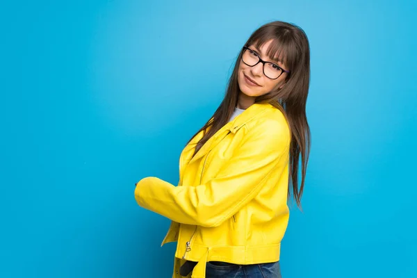 Young woman with yellow jacket on blue background pointing back with the index finger