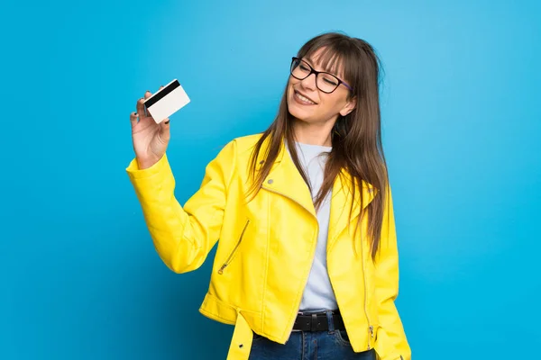 Young woman with yellow jacket on blue background holding a credit card and thinking