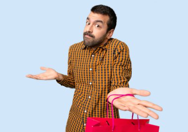 İzole mavi arka planda omuzları kaldırarak önemsiz jest yapan alışveriş çantaları ile adam