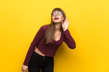 Sarı duvarın üstünde gözlüklü bir kadın. Kulağına elini sürerek bir şeyler dinliyor.