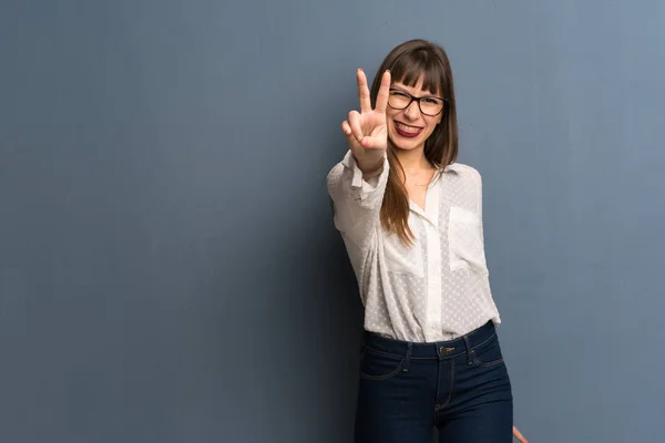 Woman with glasses over blue wall smiling and showing victory sign