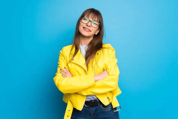 Young woman with yellow jacket on blue background keeping the arms crossed in frontal position