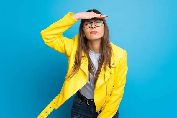 Young woman with yellow jacket on blue background looking far away with hand to look something