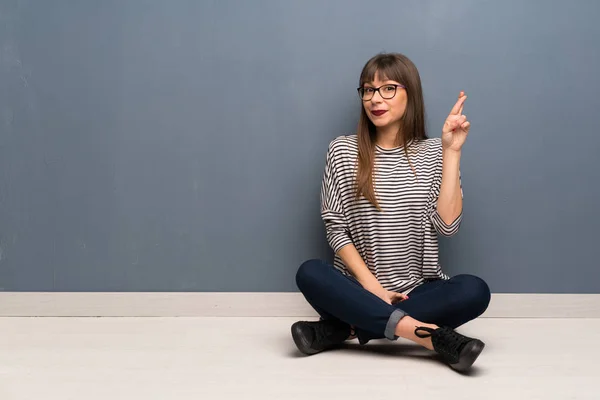Woman with glasses sitting on the floor with fingers crossing and wishing the best