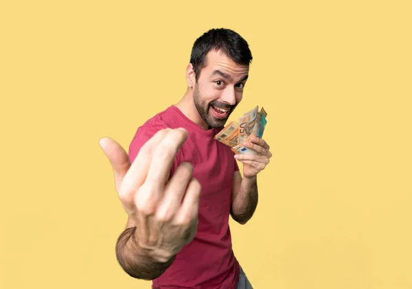 Man taking a lot of money inviting to come with hand. Happy that you came on isolated yellow background