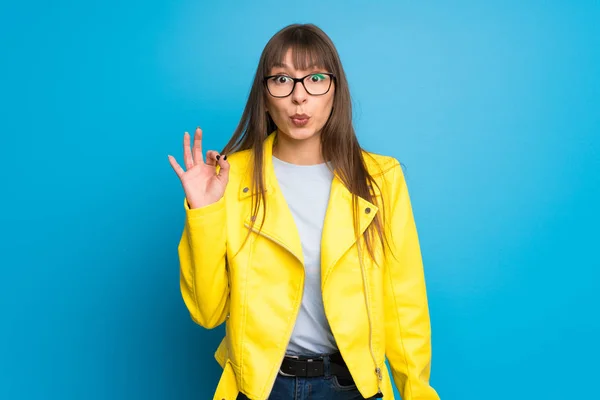 Young woman with yellow jacket on blue background surprised and showing ok sign