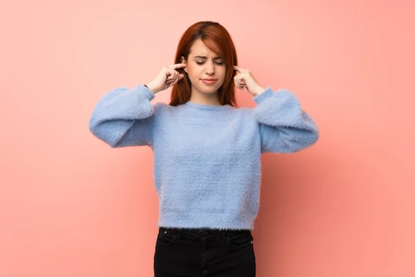 Young redhead woman over pink background covering ears with hands. Frustrated expression