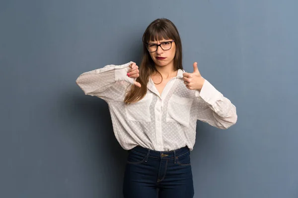 Woman with glasses over blue wall making good-bad sign. Undecided between yes or not