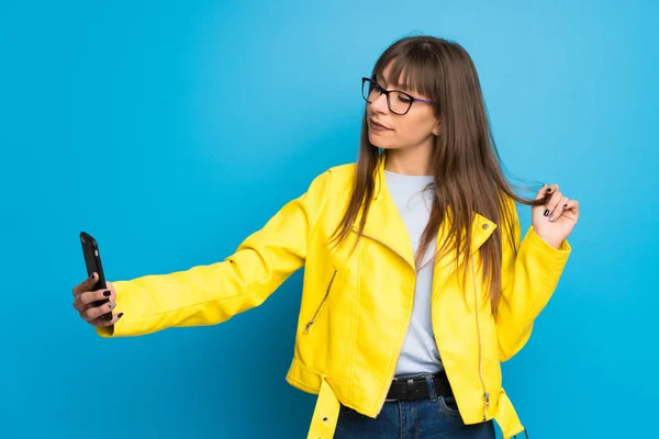 Young woman with yellow jacket on blue background making a selfie