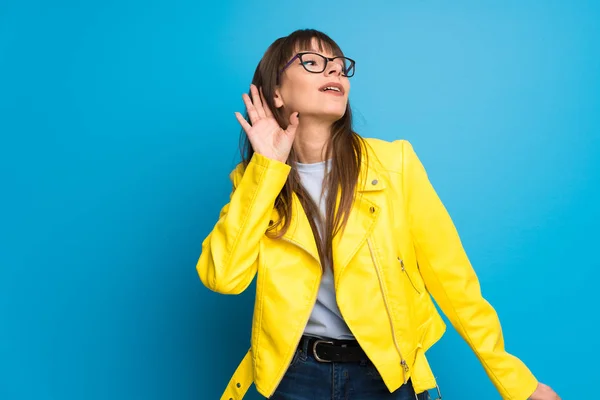 Young woman with yellow jacket on blue background listening to something by putting hand on the ear