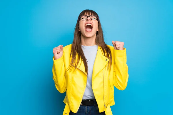 Young woman with yellow jacket on blue background shouting to the front with mouth wide open