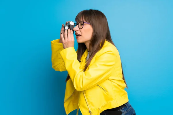 Young woman with yellow jacket on blue background holding a camera