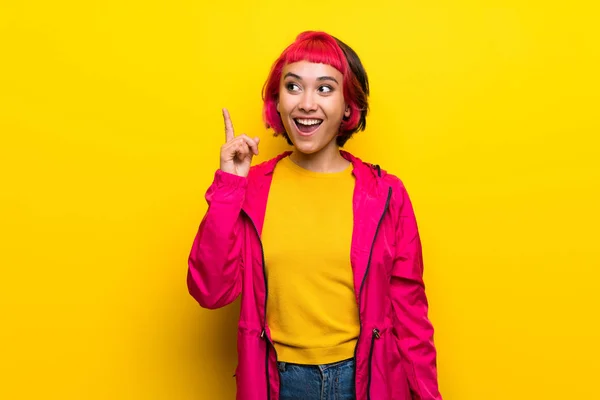 年轻的妇女与粉红色的头发在黄色的墙壁上打算实现解决方案 同时举起一个手指 — 图库照片