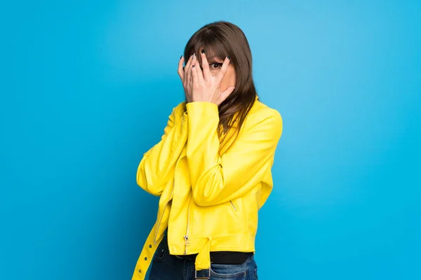 Young woman with yellow jacket on blue background covering eyes by hands and looking through the fingers