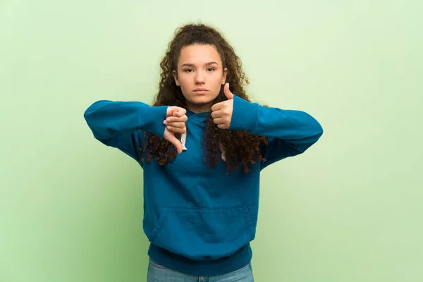 Teenager girl over green wall making good-bad sign. Undecided between yes or not