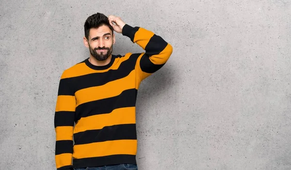 Handsome man with striped sweater having doubts while scratching head over textured wall