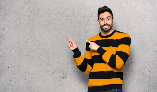 Handsome man with striped sweater frightened and pointing to the side over textured wall