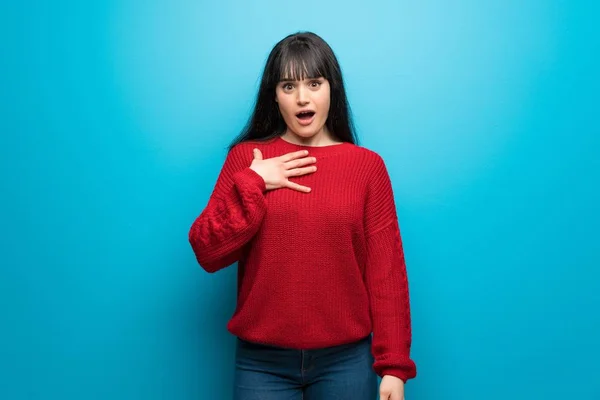 Woman with red sweater over blue wall surprised and shocked while looking right