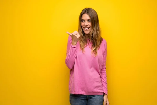 製品を提示する側を指している黄色の壁にピンクのセーターの女性 — ストック写真