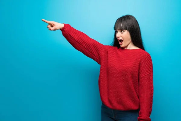 Woman with red sweater over blue wall pointing away