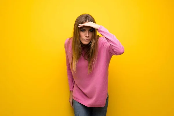 黄色い壁の上にピンクのセーターを持った女性が何かを見るために手で遠くを見ている — ストック写真