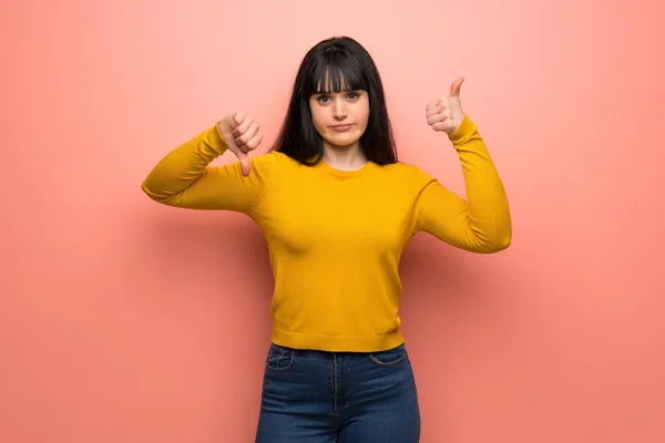 Woman with yellow sweater over pink wall making good-bad sign. Undecided between yes or not