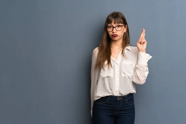 Woman with glasses over blue wall with fingers crossing and wishing the best