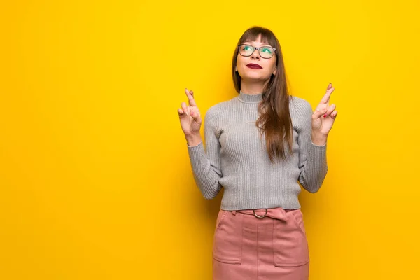 Woman with glasses over yellow wall with fingers crossing and wishing the best