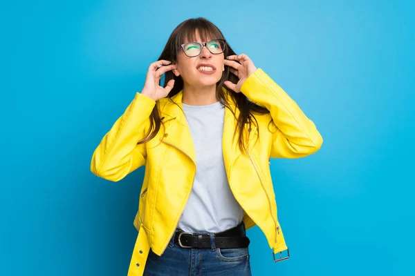 Young woman with yellow jacket on blue background covering ears with hands. Frustrated expression