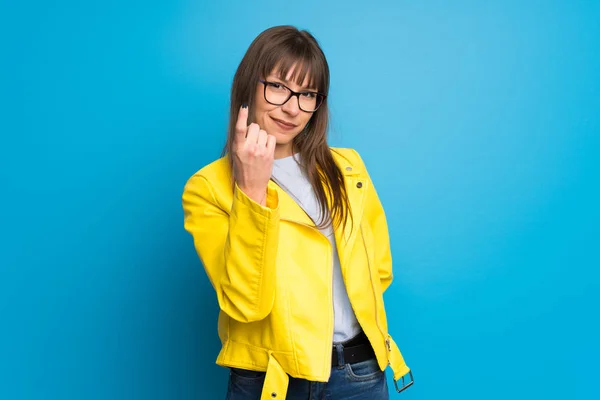 Young woman with yellow jacket on blue background inviting to come with hand. Happy that you came