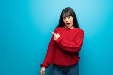 Mavi duvar sürpriz üzerinde kırmızı kazak ve işaret yan kadın ile