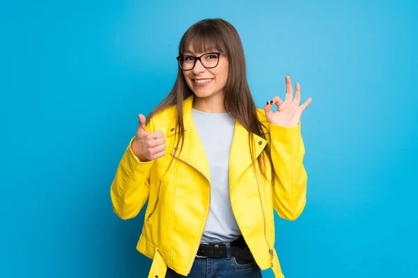 Young woman with yellow jacket on blue background showing ok sign with and giving a thumb up gesture