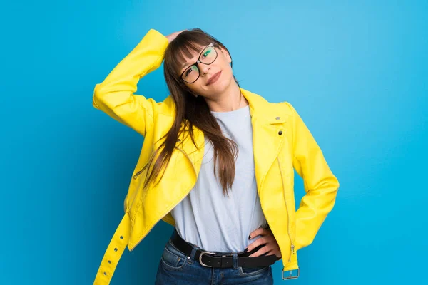 Young woman with yellow jacket on blue background thinking an idea while scratching head