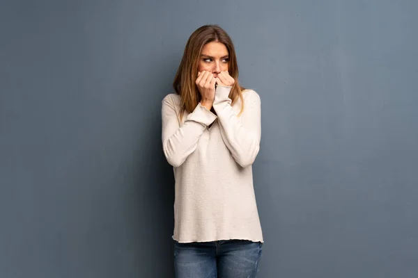 Blonde woman over grey background nervous and scared putting hands to mouth