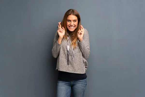 Blonde woman over grey background with fingers crossing and wishing the best