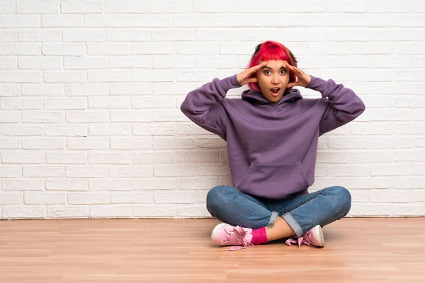 Young woman with pink hair sitting on the floor with surprise expression