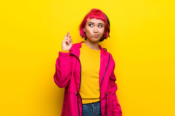 Young woman with pink hair over yellow wall with fingers crossing and wishing the best