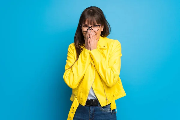 Young woman with yellow jacket on blue background smiling a lot while covering mouth
