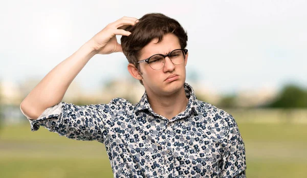 Teenager man with flower shirt and glasses having doubts while scratching head at outdoors