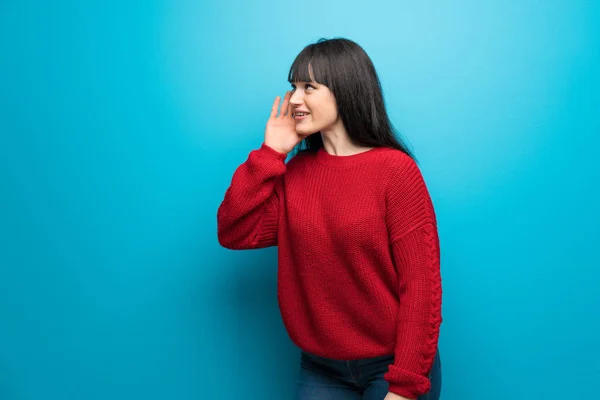 Woman with red sweater over blue wall shouting with mouth wide open to the lateral