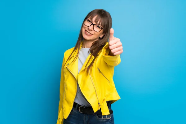 Young woman with yellow jacket on blue background giving a thumbs up gesture because something good has happened