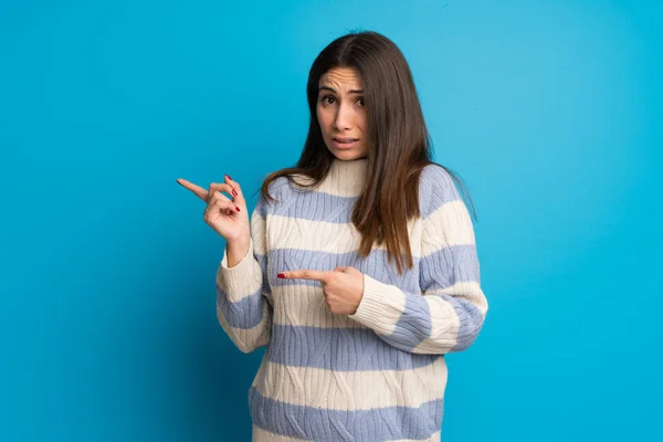 Young woman over blue wall frightened and pointing to the side