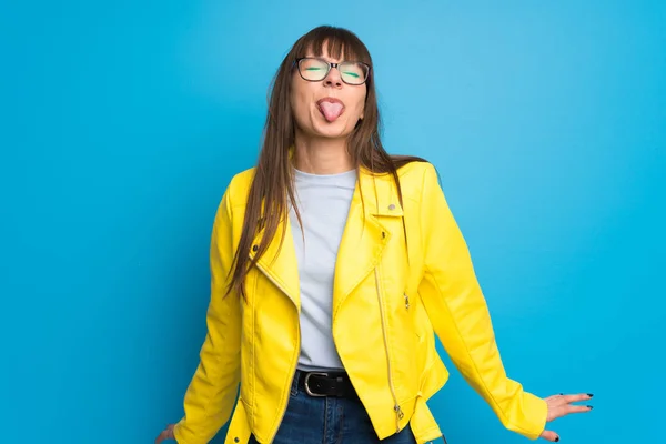 Young woman with yellow jacket on blue background showing tongue at the camera having funny look