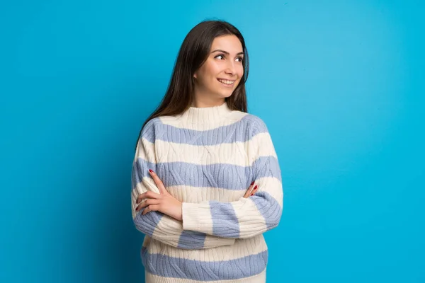 Young woman over blue wall keeping the arms crossed while smiling