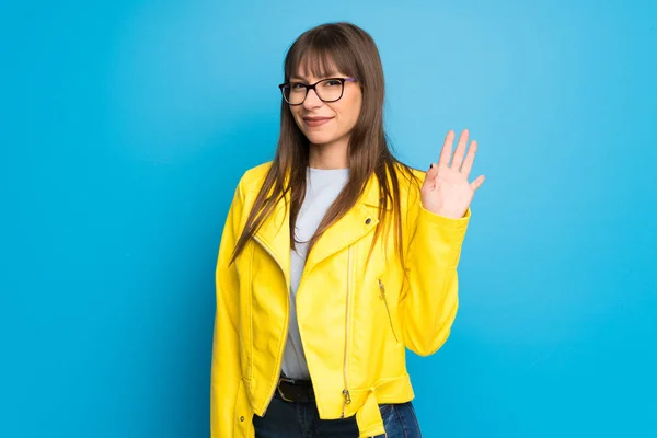 Young woman with yellow jacket on blue background saluting with hand with happy expression