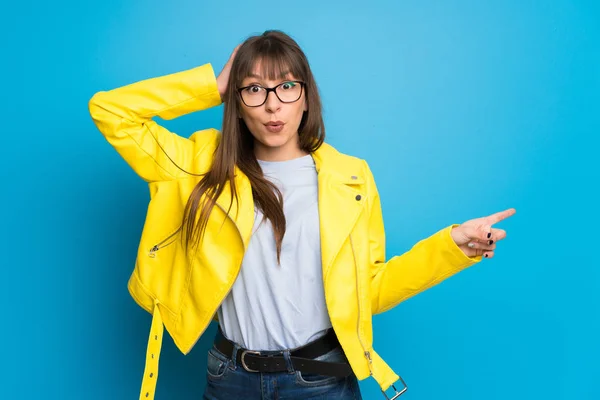 Young woman with yellow jacket on blue background pointing finger to the side and presenting a product